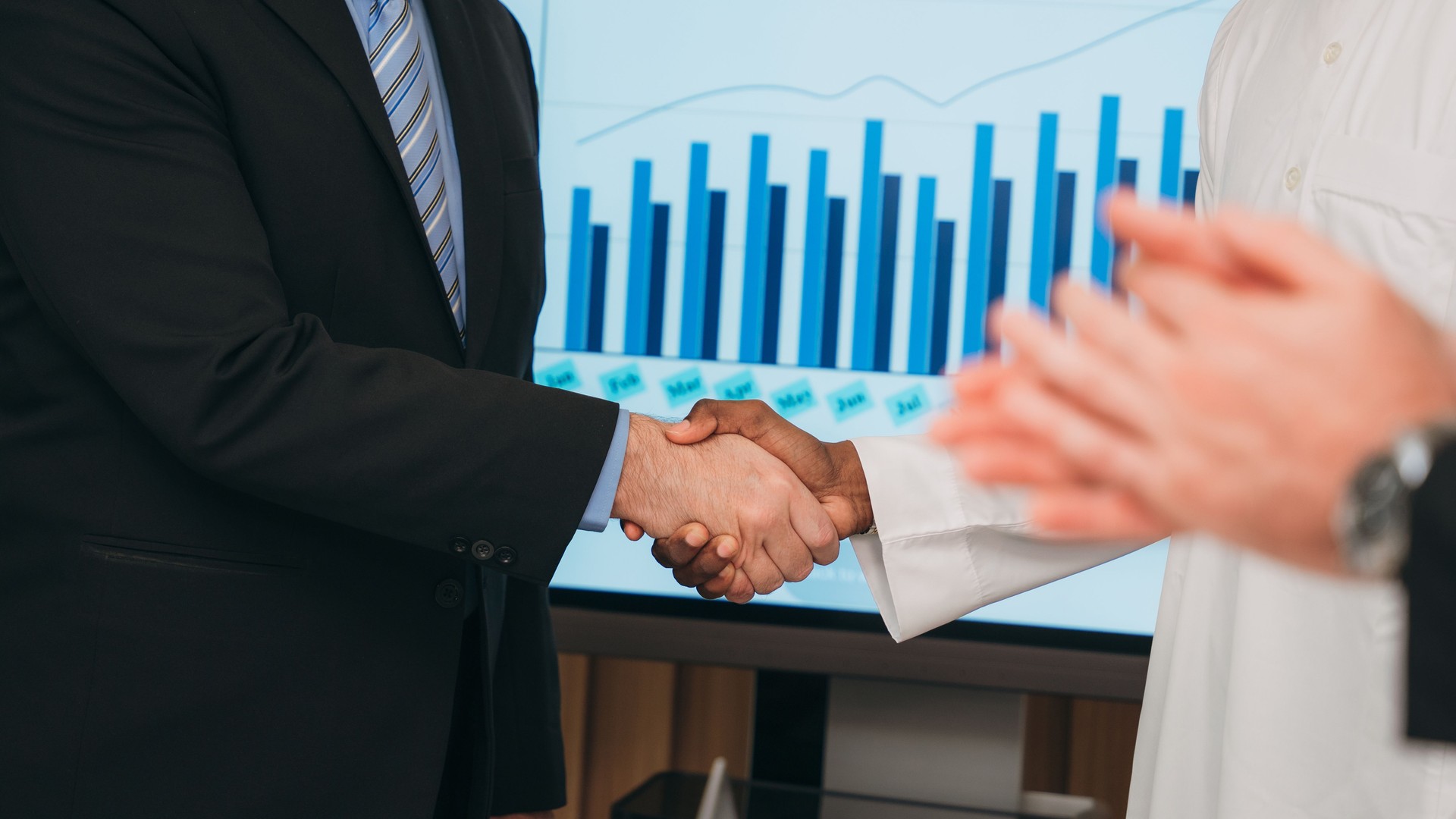 Two business men shaking hands in front of a large screen with graphs on it
