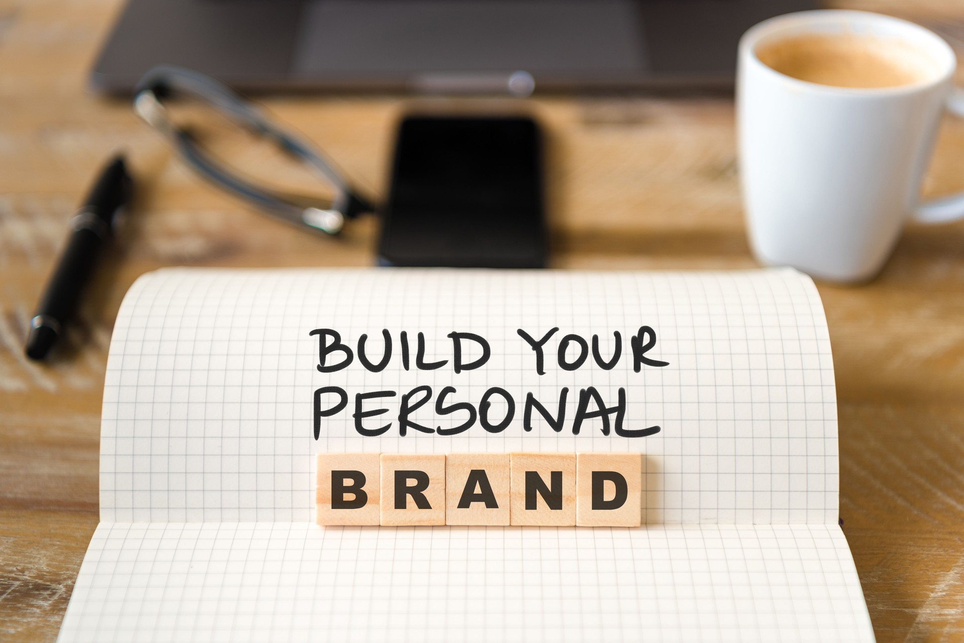 Closeup on notebook over vintage desk background, front focus on wooden blocks with letters making Build Your Personal Brand text